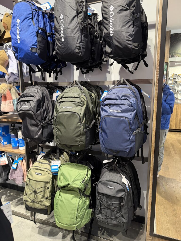 Щанд в магазин Columbia с подредени туристически и градски раници в синьо, черно и различни нюанси на зелено