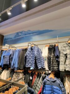 Щендери в магазин Columbia с подредени зимни якета, панталони и тениски пред голям фототапет със снежни планини