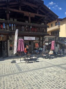 Външен изглед на кафене DABOV Specialty Coffee в Банско с традиционна дървена фасада, каменна зидария и открито пространство с маси, столове и чадъри, на които седят хора, на калдъръмена улица.