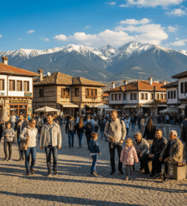 Хора от различни възрасти и националности се разхождат и почиват на павирания централен площад на Банско, заобиколен от традиционни български къщи и на фона на величествените заснежени върхове на Пирин.