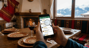 Hands holding a smartphone with a screen showing a hotel and restaurant review app with a magnifying glass icon on it. In the foreground is a rustic wooden table with traditional patterned pottery and a glass of red wine. In the background is a lit fireplace and a large window with a view of snow-capped mountains