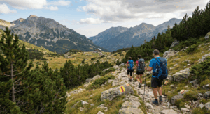 Three hikers with backpacks and poles walk along a rocky marked trail in Pirin, with a view of a mountain hut in the valley and high peaks in the background.