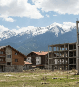 Изглед към Банско, показващ рязък контраст между голям недовършен бетонен строеж с табела и завършени ваканционни вили в алпийски стил, на фона на заснежените върхове на Пирин планина.