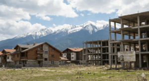 A view of Bansko, showing a stark contrast between a large unfinished concrete construction site with a sign and completed Alpine-style holiday villas, against the backdrop of the snow-capped peaks of the Pirin Mountains.