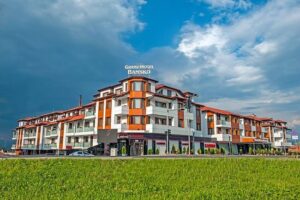 Front view of Grand Hotel Bansko with roof lettering and ground floor casino, set amidst greenery under cloudy skies in Bansko.