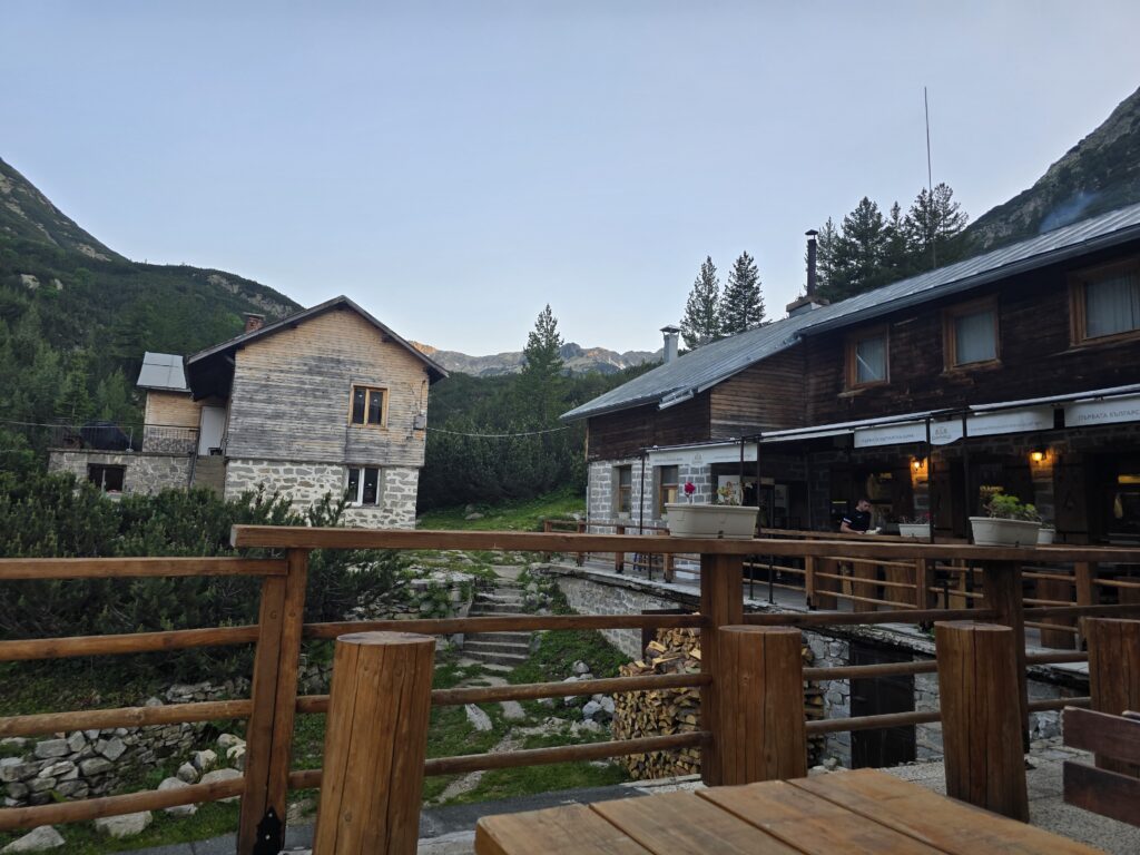 Поглед от задната дървена тераса на хижа Вихрен в Пирин, показващ двете каменни сгради на комплекса и иглолистната гора на заден план.