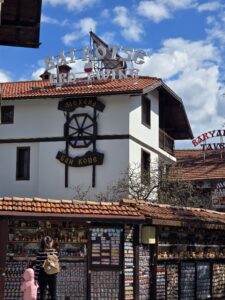 Фасада на Механа Бай Коце в Банско с голямо декоративно дървено колело и посетители, разглеждащи щанд за сувенири.