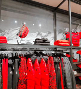 Щендер с червени и черни зимни якета, пуловери и чанти от колекцията на Moncler в магазин с планински декор на заден план.