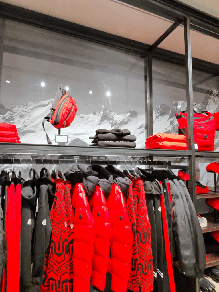 A display of red and black winter jackets, sweaters, and bags from the Moncler collection in a store with a mountain backdrop.