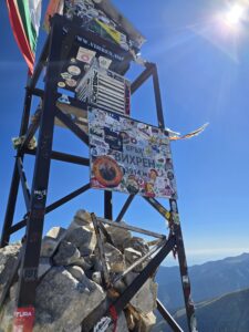 Връх Вихрен туристически панел със стикери и логота на хелиборда, със скали в подножието и планински пейзаж в позадина под ясно небе