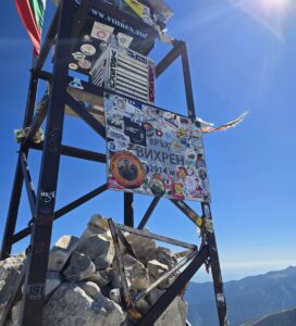 Връх Вихрен туристически панел със стикери и логота на хелиборда, със скали в подножието и планински пейзаж в позадина под ясно небе