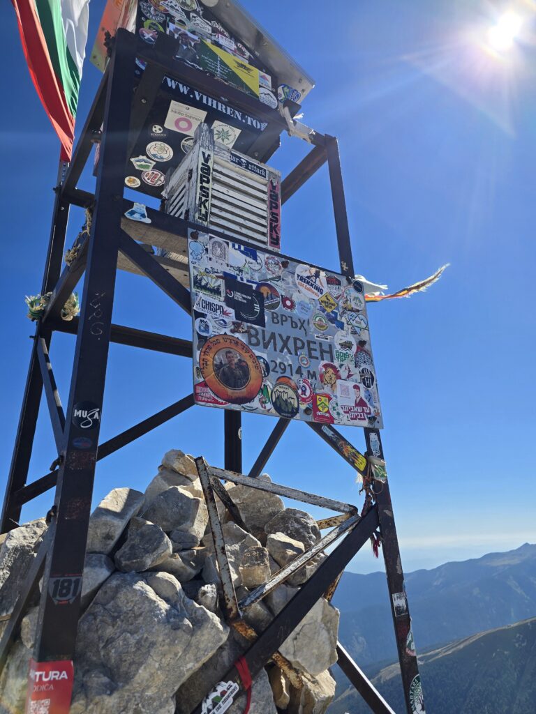 Връх Вихрен туристически панел със стикери и логота на хелиборда, със скали в подножието и планински пейзаж в позадина под ясно небе