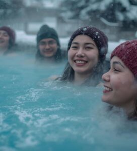 Четирима усмихнати млади хора с плетени шапки, релаксиращи в парещ открит термален басейн, заобиколен от дълбок сняг през зимата.