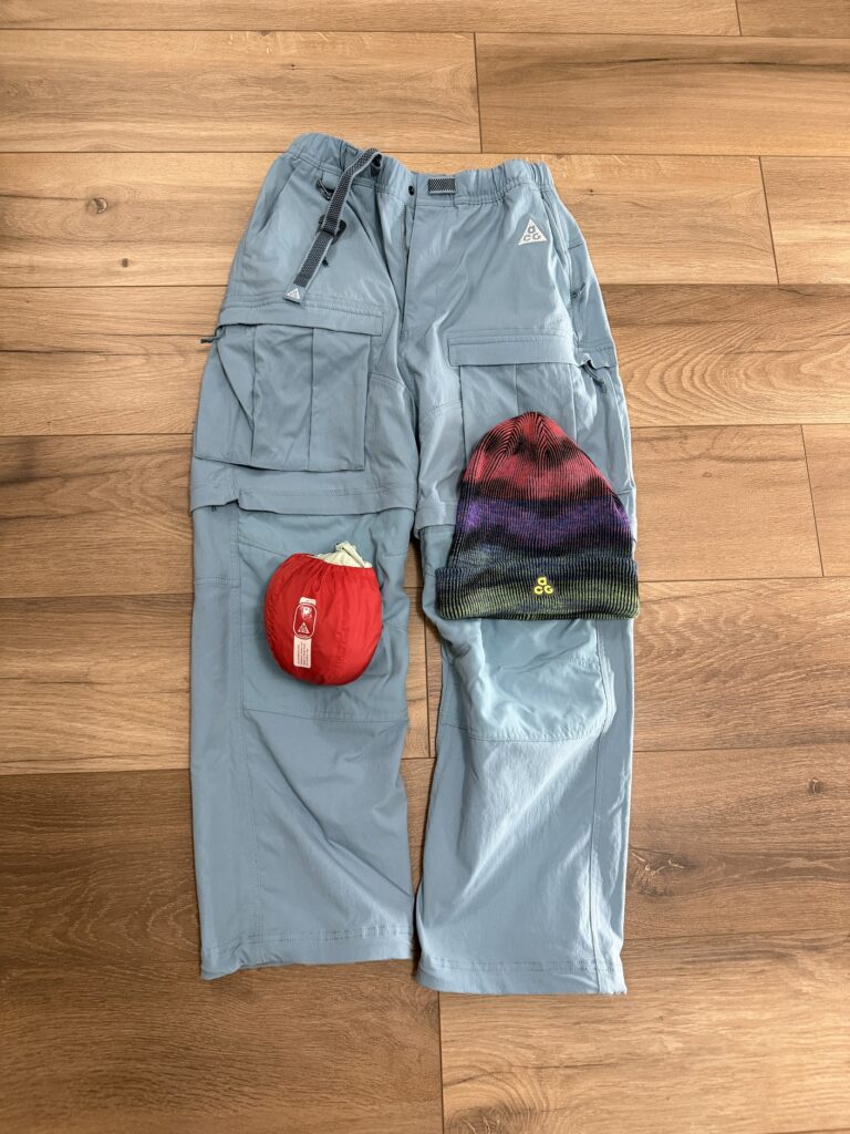 Светлосини туристически панталони с карго джобове, сгъваемо червено яке и плетена шапка в преливащи цветове, подредени върху дървен под.