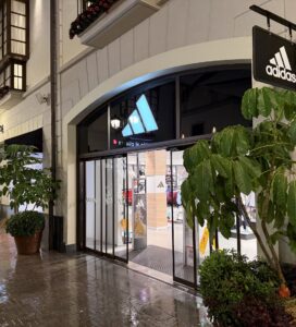Вход на магазин Adidas с осветена витрина и зелени растения отпред в търговски център вечер.