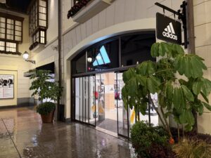 Entrance to an Adidas store with a lit window and green plants in front in a shopping mall at night.