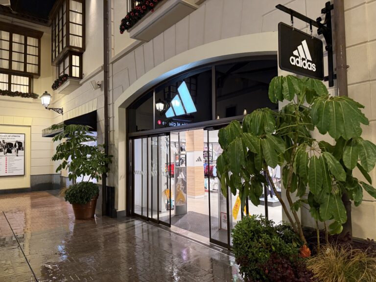 Entrance to an Adidas store with a lit window and green plants in front in a shopping mall at night.