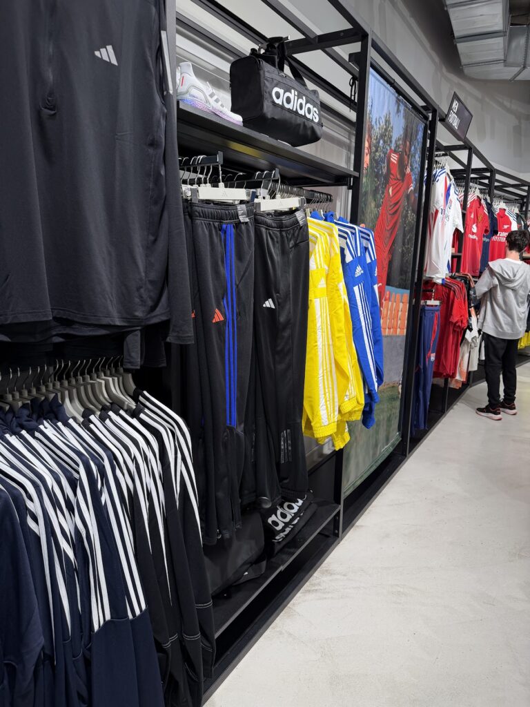 Щендери с дрехи и екипи Adidas в спортен магазин, с мъжки спортни екипи и якета в различни цветове.