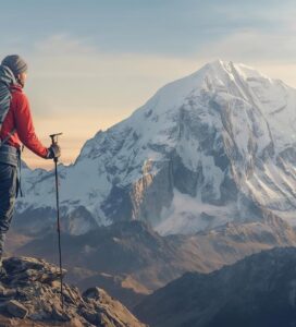 Турист с пълна екипировка, раница и щеки, стоящ на скалист ръб и гледащ към заснежения връх Вихрен в Пирин.