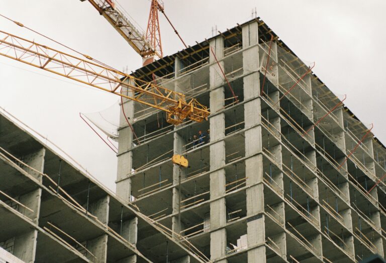 an unfinished concrete residential building in the "rough construction" stage, with a yellow construction crane and safety net, against a gloomy, gray sky