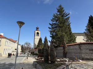 Изглед към емблематичната каменна часовникова кула и църквата "Света Троица" зад каменен зид в Банско, разположени до слънчева пешеходна улица с разхождащи се хора.