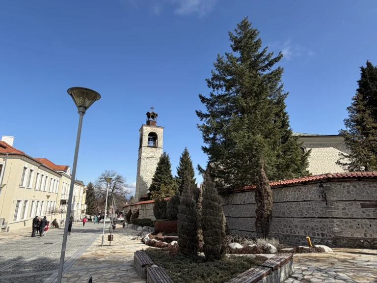 Изглед към емблематичната каменна часовникова кула и църквата "Света Троица" зад каменен зид в Банско, разположени до слънчева пешеходна улица с разхождащи се хора.