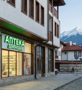 Денонощна аптека в Банско с голям зелен светещ кръст и надпис "АПТЕКА ДЕНОНОЩНА". Сградата е на калдъръмена улица, а на заден план се виждат заснежените върхове на Пирин планина.