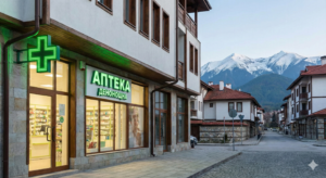 A 24-hour pharmacy in Bansko with a large green illuminated cross and the inscription "24-hour pharmacy". The building is on a cobblestone street, and in the background the snow-capped peaks of Pirin Mountain are visible.