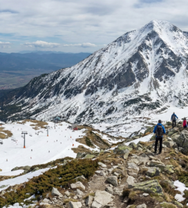 Зимен пейзаж на връх Тодорка, извисяващ се над ски зона Банско с работещи лифтове. На преден план се виждат туристи на скалист гребен, а в долината – град Банско.