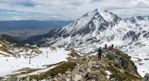 Winter landscape of Todorka Peak, towering above the Bansko ski area with working lifts. In the foreground, tourists are seen on a rocky ridge, and in the valley - the town of Bansko.