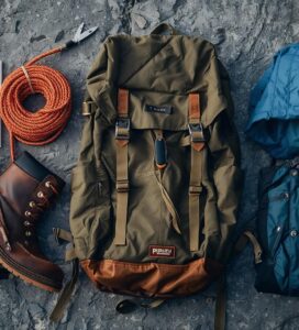 Колекция от планинска екипировка – туристически обувки, раница, яке и катерачен инвентар, подредени върху скалиста повърхност.