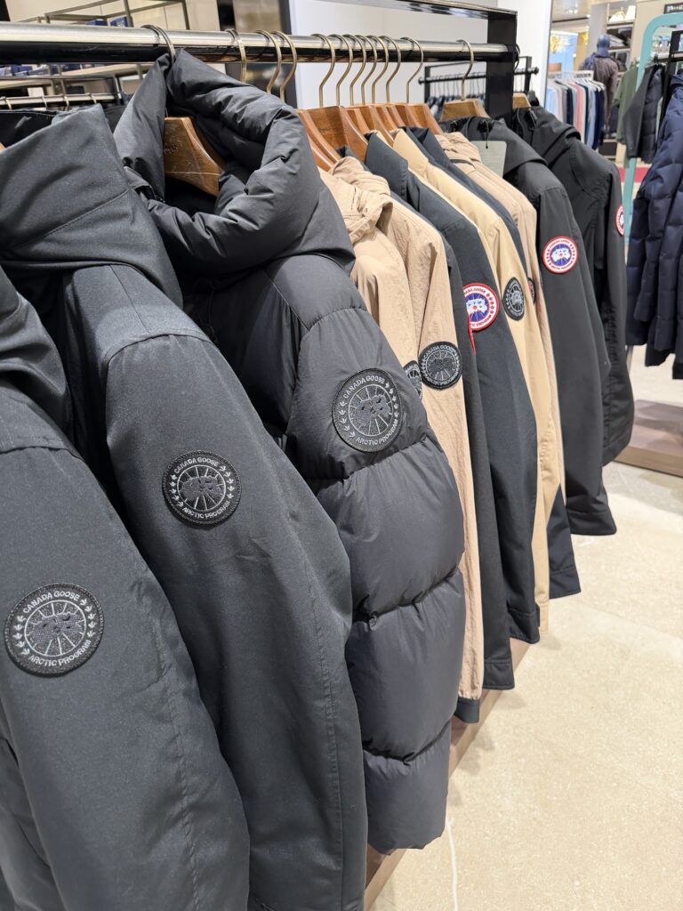 Ред от зимни якета Canada Goose в черно и бежово, подредени на закачалки в магазин, с видими кръгли нашивки с логото на марката.
