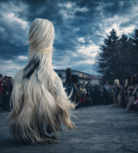 Кукери с традиционни кожени костюми танцуват на площада, обградени от публика, под драматично облачно небе