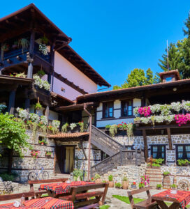 Снимка на традиционна българска къща за гости или механа, изградена от камък и тъмно дърво, с множество саксии с цветя по балконите и дървени маси с червени карирани покривки в слънчевия двор.