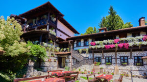 A photo of a traditional Bulgarian guesthouse or tavern, built of stone and dark wood, with numerous flower pots on the balconies and wooden tables with red checkered tablecloths in the sunny courtyard.