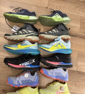 Различни модели маратонки за планина и бягане от Nike, Salomon и Adidas Terrex, подредени в редици върху дървен под.