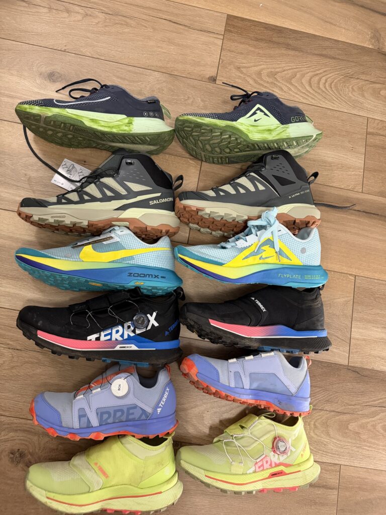 Различни модели маратонки за планина и бягане от Nike, Salomon и Adidas Terrex, подредени в редици върху дървен под.