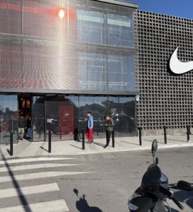 Фасада на модерен магазин Nike с голямо бяло лого и стъклен вход, пред който минават хора в слънчев ден.
