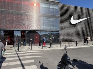 Facade of a modern Nike store with a large white logo and a glass entrance, with people walking past on a sunny day.