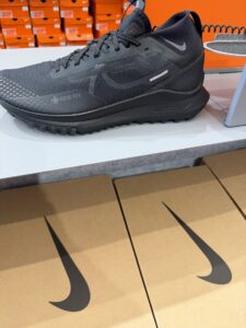 Черни маратонки Nike Gore‑Tex за трейл бягане, изложени върху плот, с кафяви кутии с логото Swoosh на преден план.