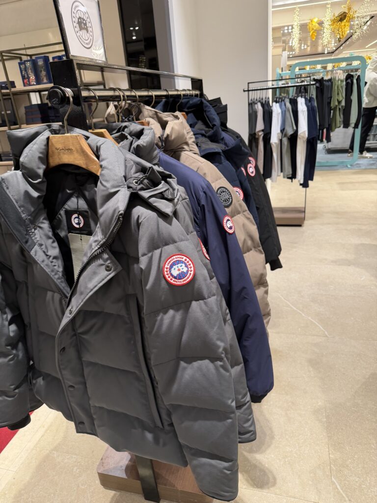 Ред от зимни якета Canada Goose в сиво, бежово и тъмносиньо, подредени на закачалки в магазин, с ясно видими кръгли емблеми на ръкавите.