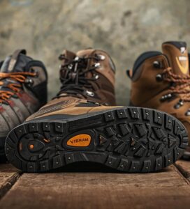 Три броя високи туристически обувки (hiking boots) върху дървена повърхност на фона на карта. Централната обувка е обърната настрани, показвайки детайлна черна гумена подметка с оранжев надпис "VIBRAM". Другите две обувки са изправени, в кафяви и сиви нюанси, с връзки.
