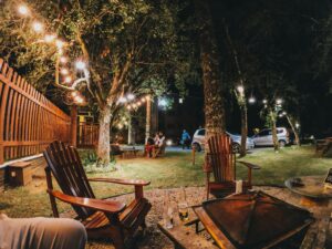 Night shot of a cozy backyard with wooden chairs, a fire pit, a table with beer bottles, and trees decorated with glowing garlands. People are visible in the background.