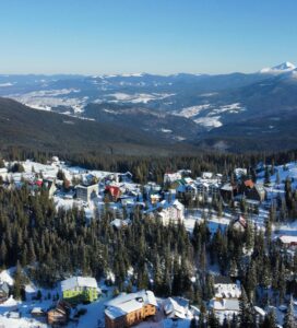 зимен курорт с множество хотели и хижи, разположени сред иглолистна гора, на фона на високи заснежени планински върхове и ясно синьо небе