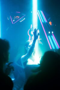 A young woman dressed in black dances with her hand raised under a bright blue spotlight in a nightclub. Behind her, neon lights and geometric elements create a party atmosphere