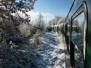 Winter landscape shot from a moving train. A dark green train carriage moves through a deep snow-covered forest under a bright blue sky.