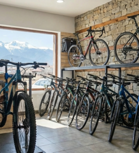 Модерен велосипеден магазин с каменна стена и дървени елементи. На преден план механик клекнал до стойка ремонтира синьо планинско колело. Вдясно са подредени редици нови велосипеди, а на заден план през голям витринен прозорец се виждат заснежените върхове на планината и синьо небе.