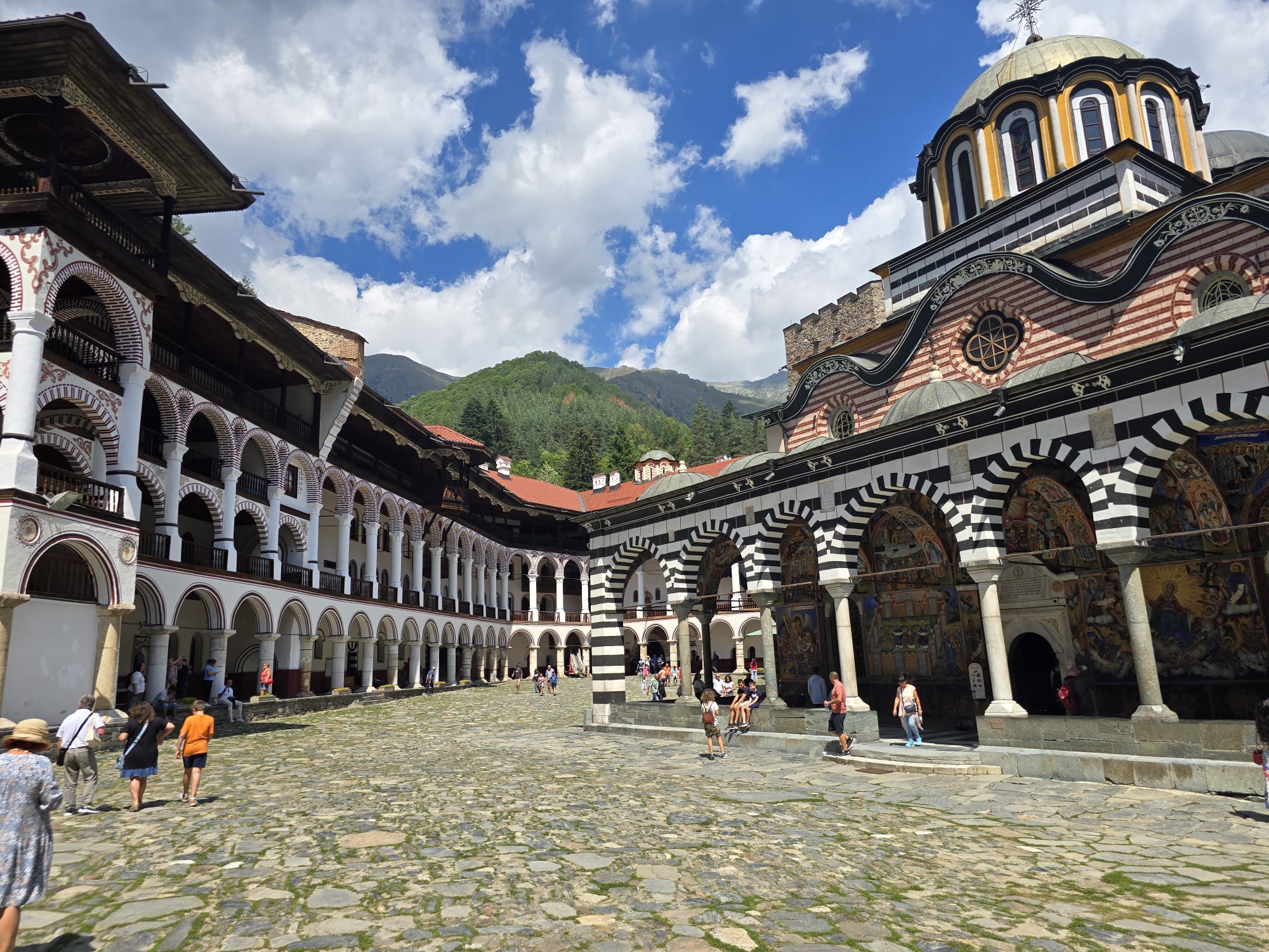 Вътрешният двор на Рилския манастир с тройните аркади на жилищните крила и централната църква с купол и боядисани черно-бели арки в слънчев летен ден