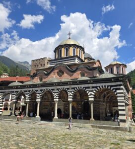 Централната църква на Рилския манастир с аркадните колонади и богато изрисувани стенописи, заобиколена от планински склонове и синьо небе с облаци