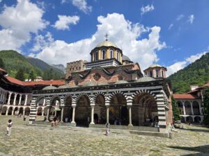 The central church of the Rila Monastery with its arcaded colonnades and richly painted frescoes, surrounded by mountain slopes and a blue sky with clouds